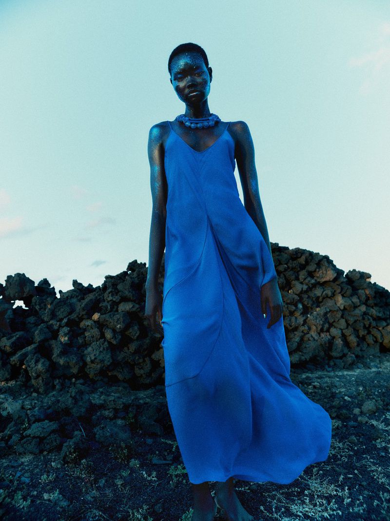 Model Ateta Jok wearing a long blue dress against a dark rocky terrain and pale sky for Sèduction Magazine.