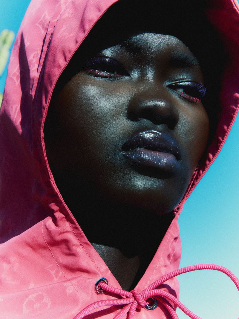 Close-up portrait of model Ateta Jok wearing a pink hooded windbreaker against a clear blue sky.