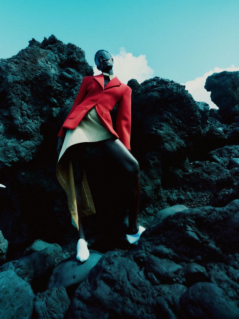 Model Ateta Jok posing on dark volcanic rock in a red structured blazer, draped beige skirt, and white pointed shoes under a blue sky.