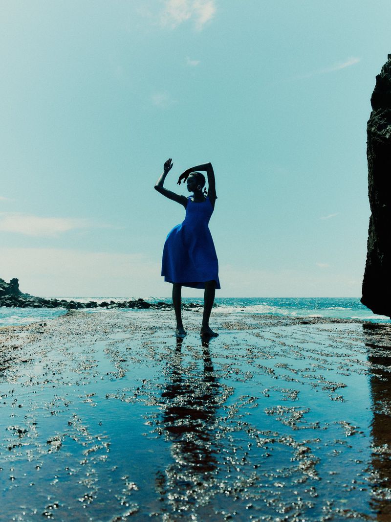 Model Ateta Jok wearing a vibrant blue dress posing gracefully in shallow coastal water against a bright blue sky.