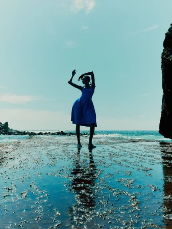 Model Ateta Jok wearing a vibrant blue dress posing gracefully in shallow coastal water against a bright blue sky.