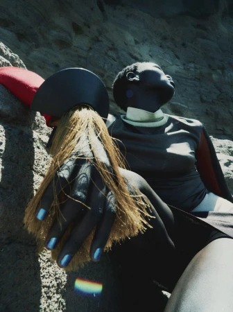 Model resting against a rocky cliff face wearing a dark top with red accents and holding a brush with long blonde bristles.
