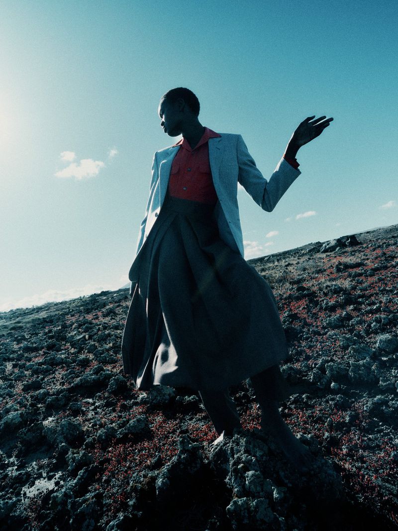 Model standing on rocky terrain wearing a grey blazer, red shirt, and dark skirt against a bright blue sky.
