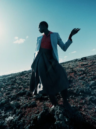 Model standing on rocky terrain wearing a grey blazer, red shirt, and dark skirt against a bright blue sky.