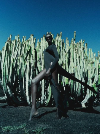 Model Ateta Jok poses in a black high-slit dress and sunglasses against a wall of towering cacti under a deep blue sky.