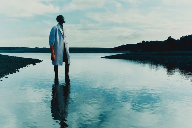 Model standing ankle-deep in calm water wearing a pale coat and orange gloves against a cloudy sky horizon.