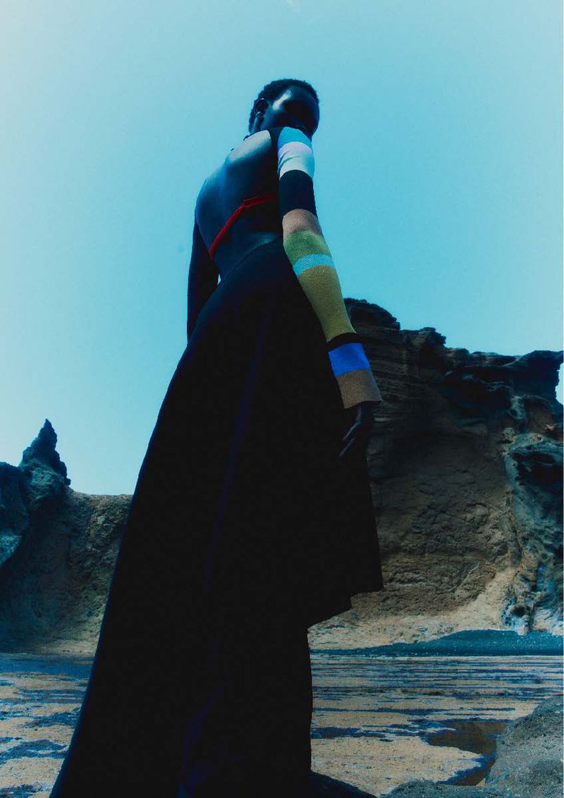 Low-angle view of model Ateta Jok wearing a black skirt and color-block sleeve top against a rocky landscape and blue sky.