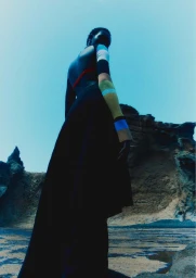 Low-angle view of model Ateta Jok wearing a black skirt and color-block sleeve top against a rocky landscape and blue sky.