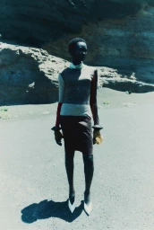 Model Ateta Jok standing outdoors in a high-neck colorblock knit top and dark skirt against rocky terrain.