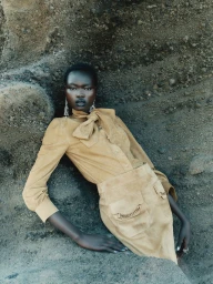 Model Ateta Jok wearing a camel suede bow-neck top and matching skirt, leaning against a rough rock wall for Sèduction Magazine.