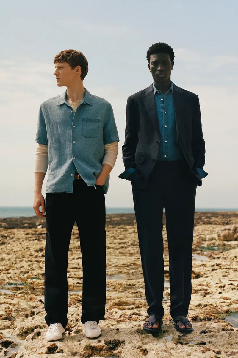 Two male models posing on a rocky coastline in Zara Origins menswear including a blue shirt, tailored suit jacket, and trousers.