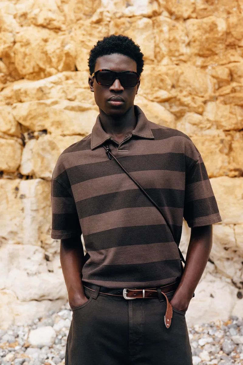 Model Cheikh Amar Diaw wearing a brown striped polo shirt and dark sunglasses against a natural rock cliff wall.