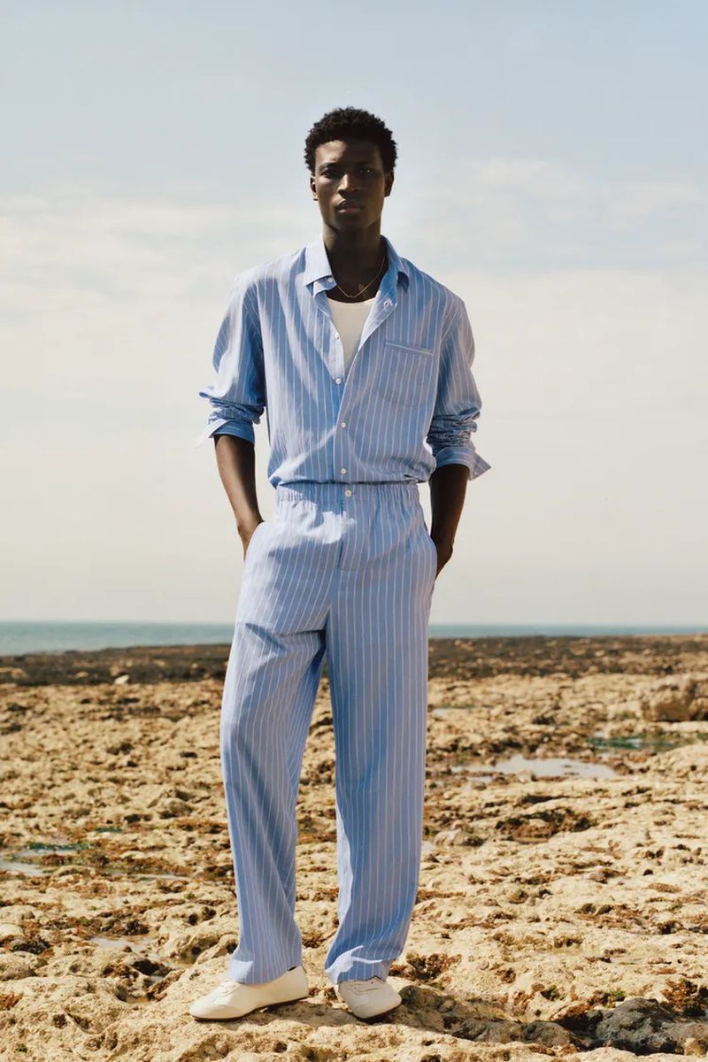 Model Cheikh Amar Diaw wearing a light blue striped matching shirt and trousers standing on a rocky coast against the ocean.