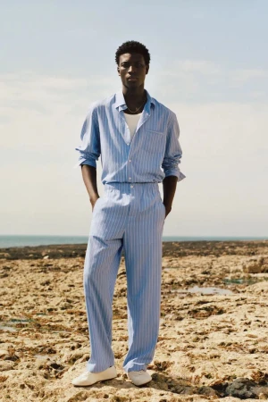 Model Cheikh Amar Diaw wearing a light blue striped matching shirt and trousers standing on a rocky coast against the ocean.