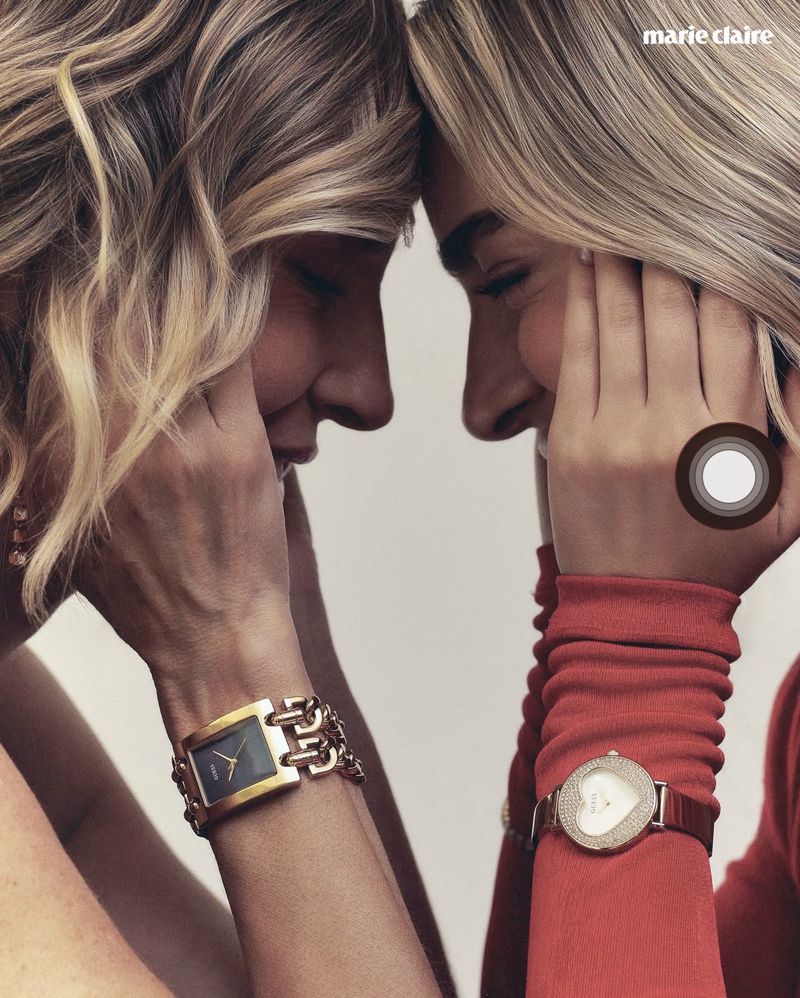 Two blonde women smiling with their foreheads touching, displaying gold and jeweled wristwatches against a light background.