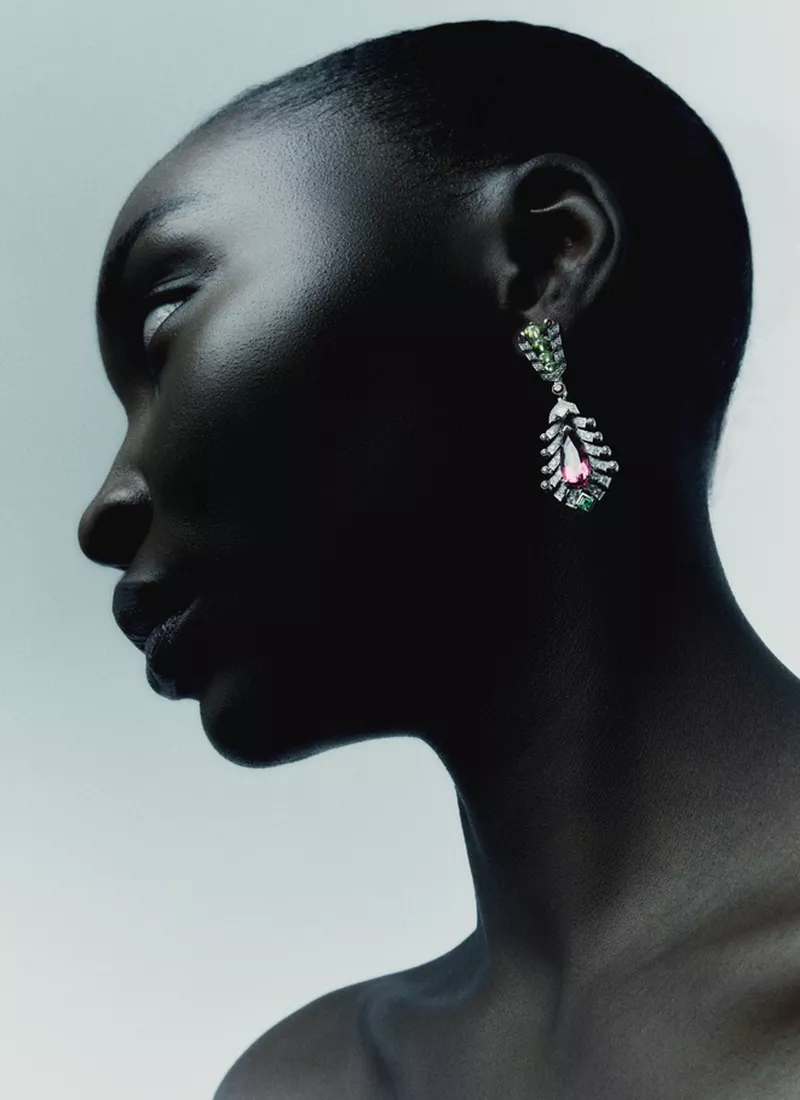 Profile portrait of a Black woman with short hair wearing an ornate gemstone drop earring against a pale gradient background.
