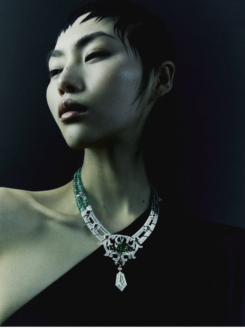 Model Wawa Li wearing an elaborate diamond and emerald statement necklace against a dark background.