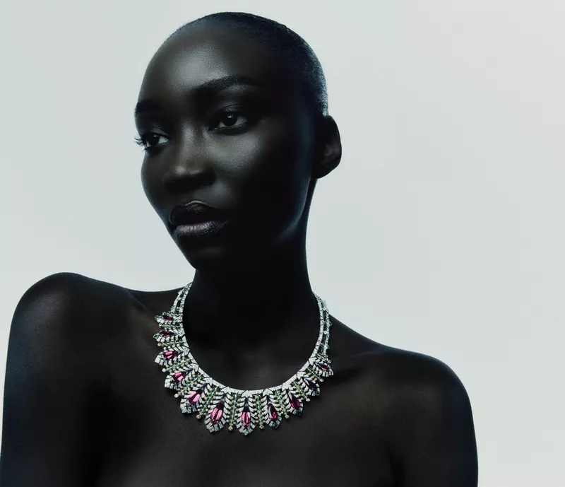 Model Diarra Ndiaye poses with bare shoulders wearing an elaborate diamond and pink gemstone collar necklace against a light studio backdrop.