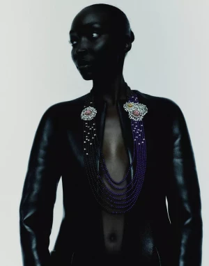 Black model with shaved head wearing an elaborate multi-strand beaded necklace with floral brooches and an open black jacket.