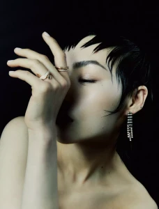 Close-up beauty portrait of model Wawa Li wearing fine rings and a dangling earring against a solid black background.