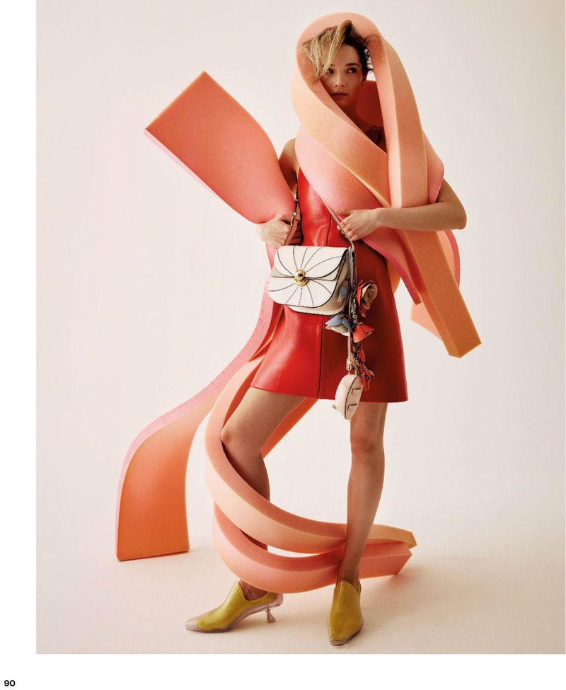 Model wearing a red mini dress and yellow heels, draped in large peach sculptural foam ribbons while holding a white handbag.