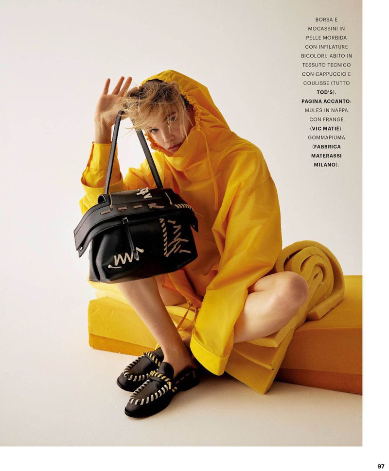 Model in a bright yellow hooded top holding a black leather handbag and wearing black loafers against a seamless white background