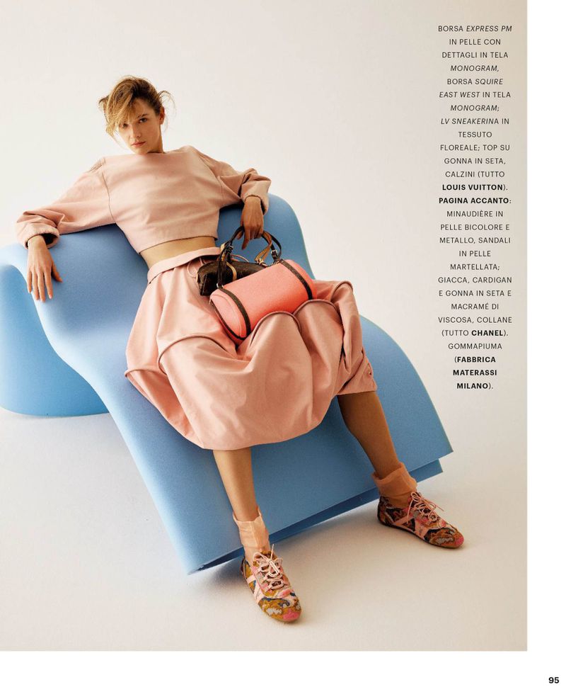 Fashion model in a pink crop top and voluminous bubble skirt seated on a curved blue foam chair against a white background.
