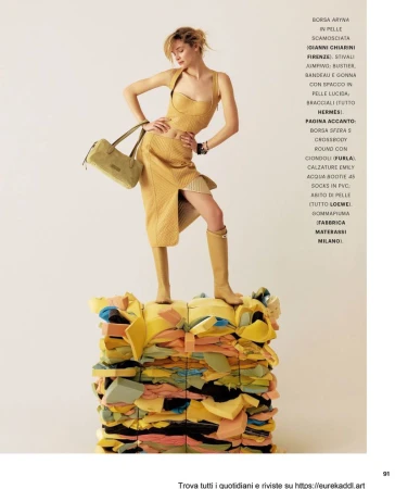 Fashion model in a textured yellow top, slit skirt, and tall boots posing on a bale of foam against a pale backdrop.