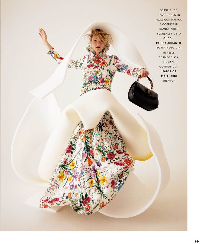 Model posing in a floral gown enveloped by a sculptural white foam structure, holding a black handbag against a pale studio backdrop.