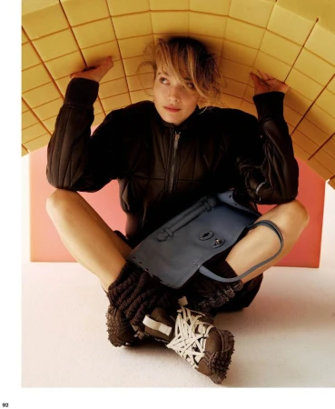 Model sitting cross-legged under a curved yellow grid structure wearing a black jacket with a blue handbag on her lap.