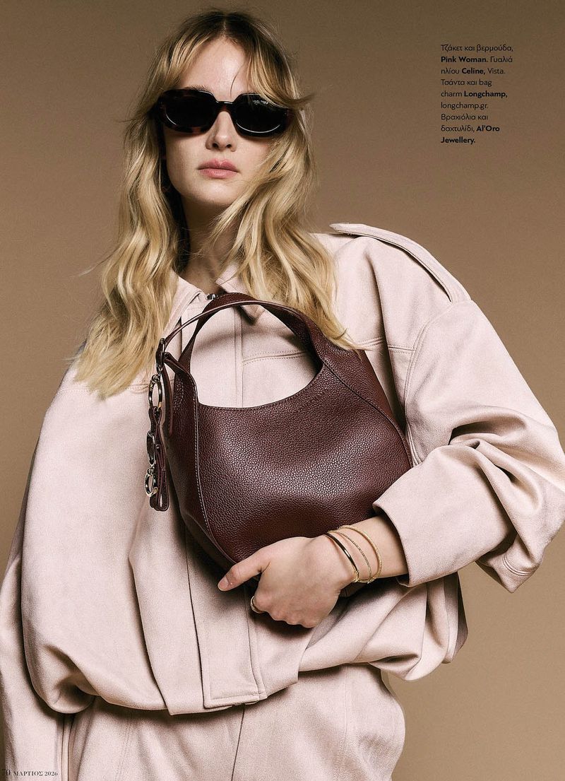 Blonde model in dark sunglasses and an oversized pale pink jacket holding a deep burgundy leather handbag against a tan studio backdrop.