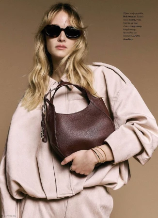 Blonde model in dark sunglasses and an oversized pale pink jacket holding a deep burgundy leather handbag against a tan studio backdrop.