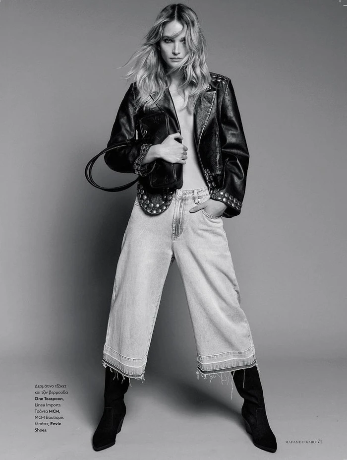 Model in an open studded leather jacket, wide-leg cropped jeans, and black boots posing against a studio backdrop.