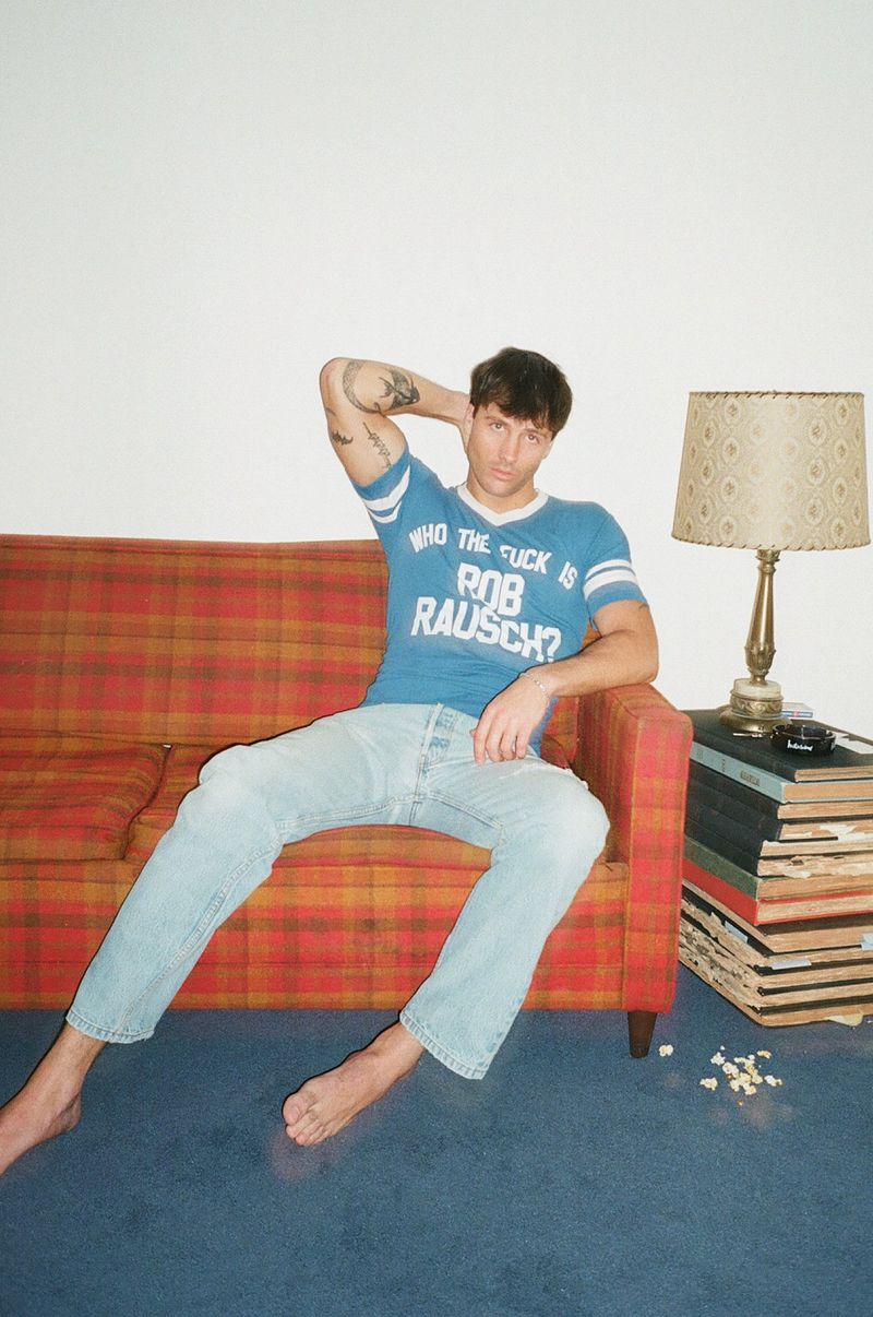 Rob Rausch sitting barefoot on a red plaid couch wearing a blue graphic t-shirt and light blue jeans.