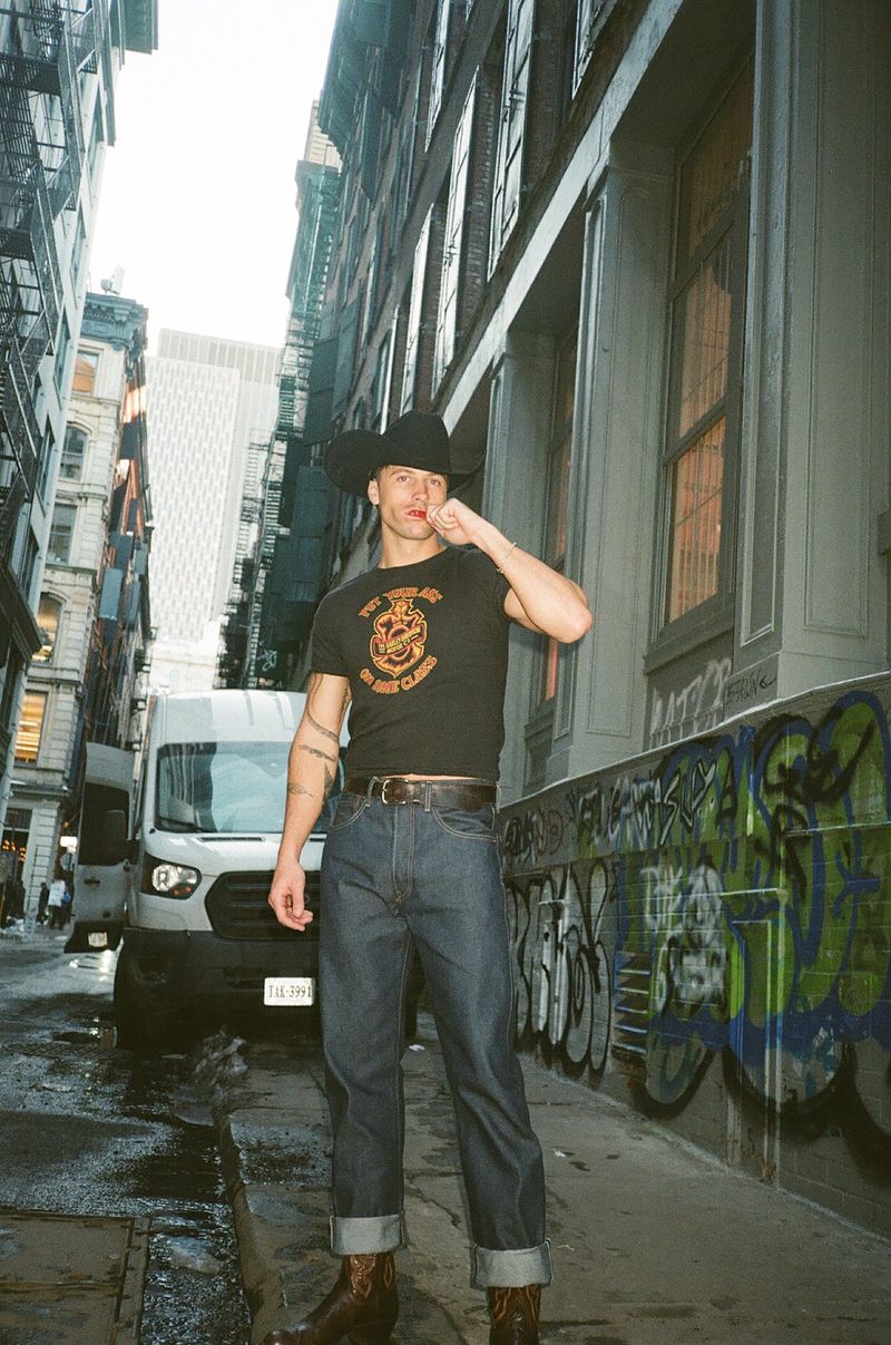 Rob Rausch wearing a black cowboy hat, graphic t-shirt, cuffed blue denim jeans, and boots in an urban city alley.