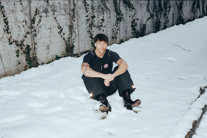 Rob Rausch seated in snow wearing a black short-sleeve top and dark trousers against a concrete wall covered in ivy.
