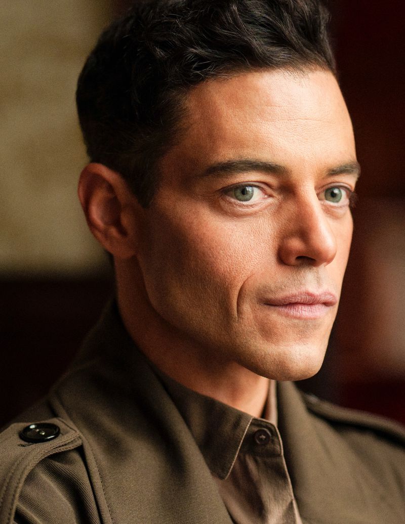 Portrait of Rami Malek in a khaki-colored jacket against a blurred neutral background, showcasing a poised and serious demeanor.