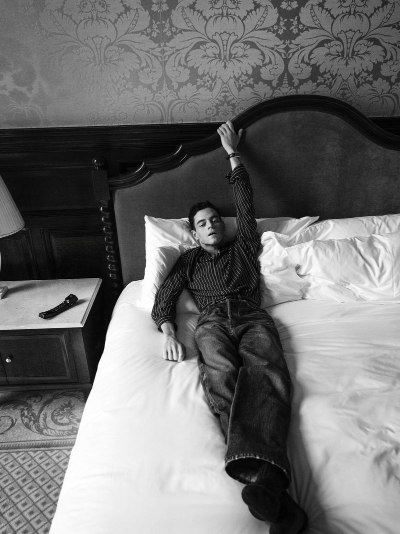 Black and white portrait of a male adult lying on a bed in casual clothes, with decorative patterned wallpaper in the background.