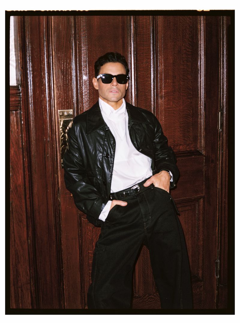 Man in leather jacket posing against wooden door. Dark sunglasses, casual shirt, black pants. Indoor fashion portrait with dramatic lighting.