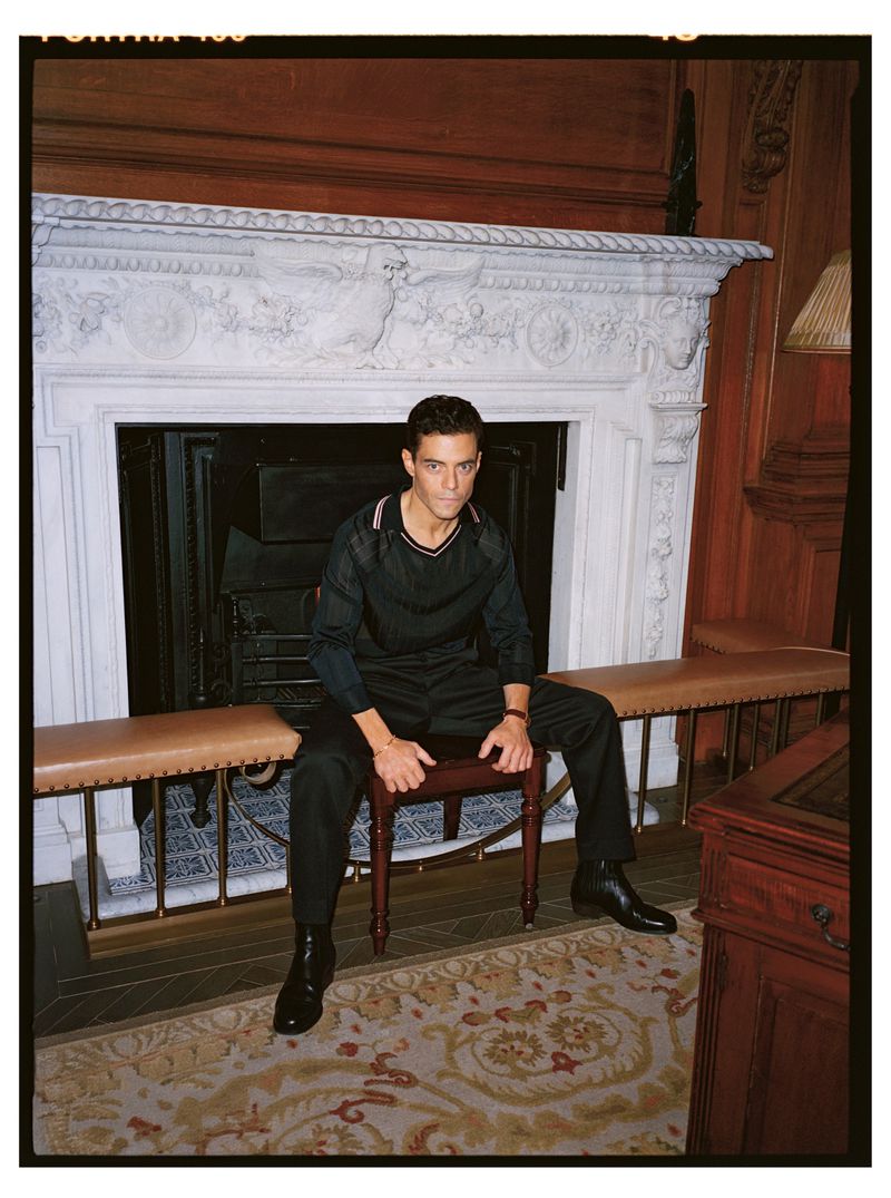 Portrait of a man in a black outfit sitting on a stool in front of a white fireplace, with wood paneling and patterned carpet.