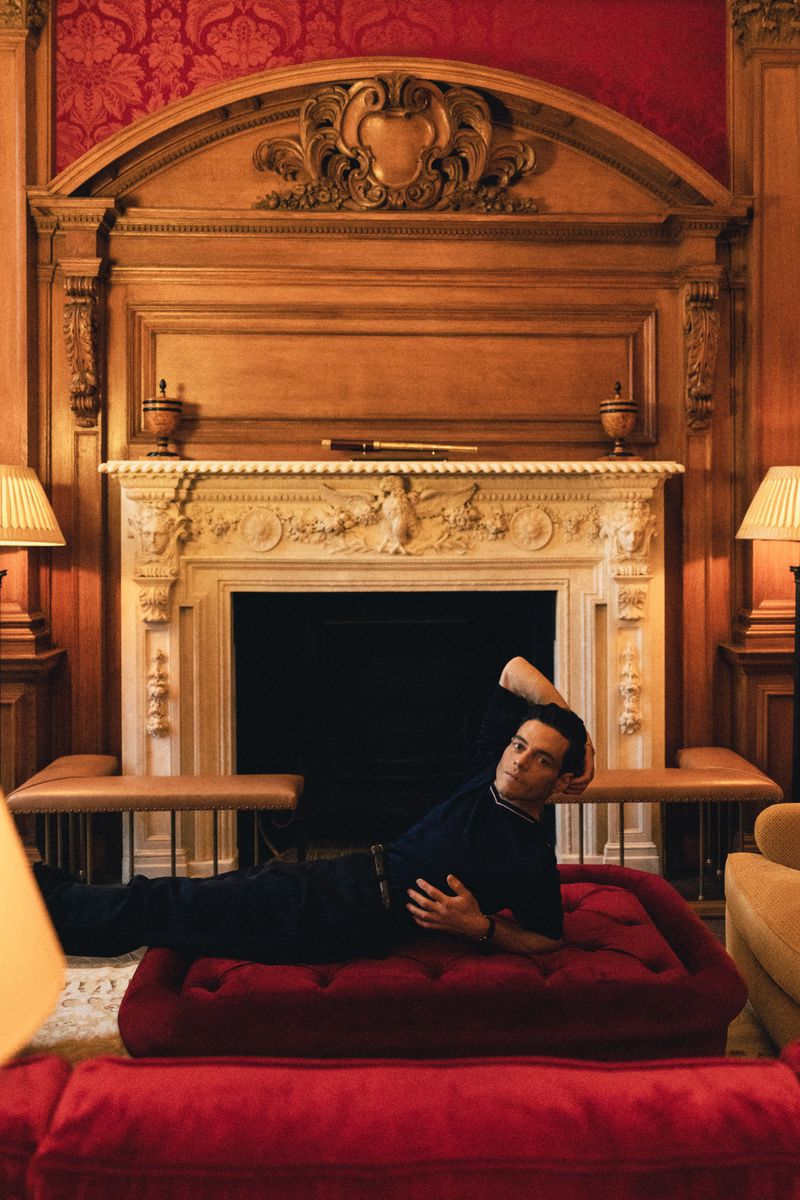 A man in a polo shirt is reclining on a red ottoman in an elegant room with paneled walls and a decorative fireplace mantel.