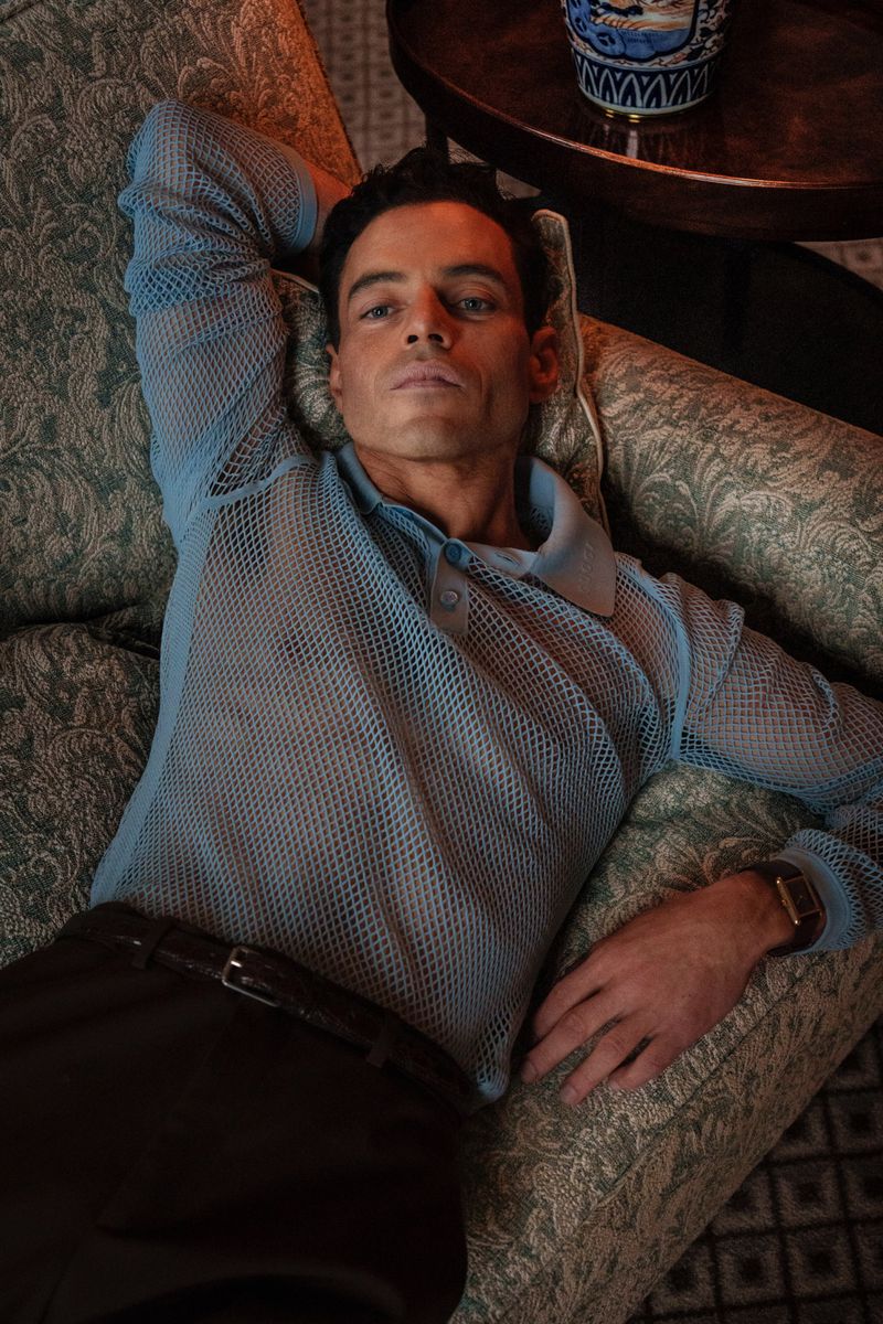 Portrait of Rami Malek in a light blue mesh shirt, lying on an ornate patterned couch in an elegant room with dark wood accents.