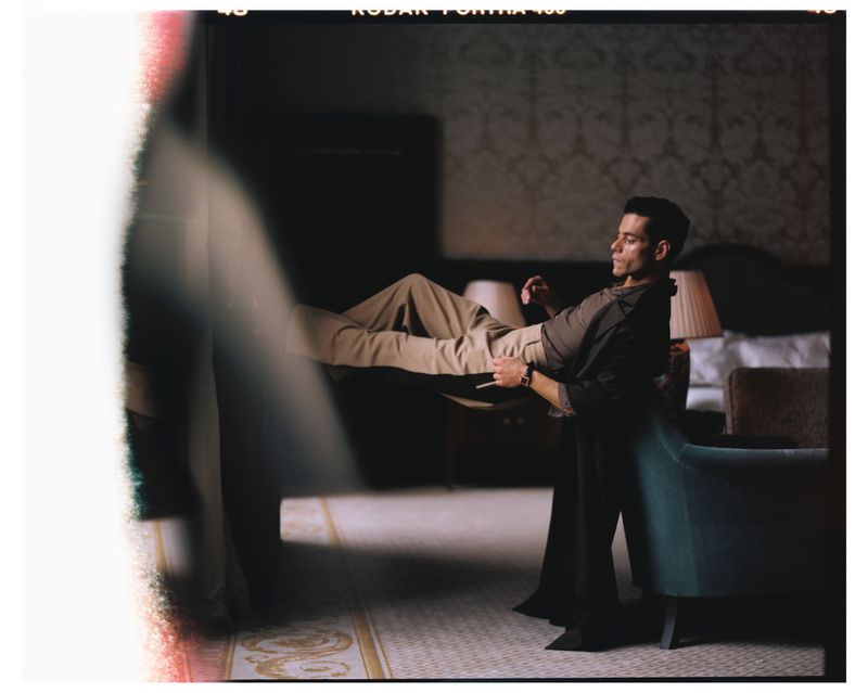 Portrait of an adult male being held in the air in a room with patterned wallpaper and a bed, lit by a lamp.