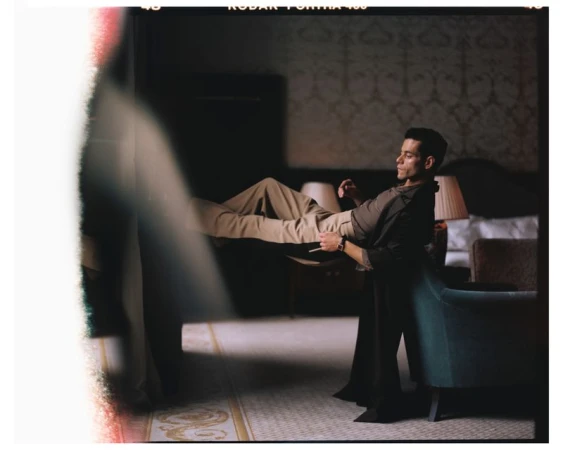Portrait of an adult male being held in the air in a room with patterned wallpaper and a bed, lit by a lamp.