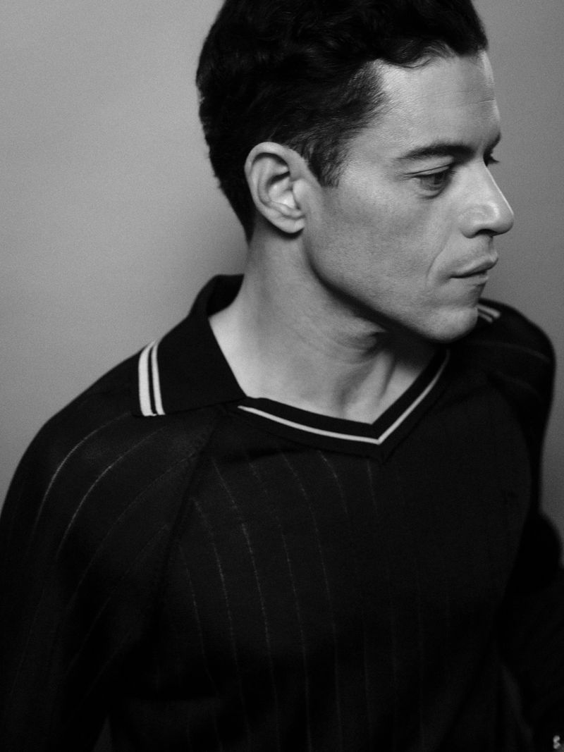 Black and white portrait of a man with dark hair, wearing a black and white striped V-neck shirt, gazing away in a studio setting.