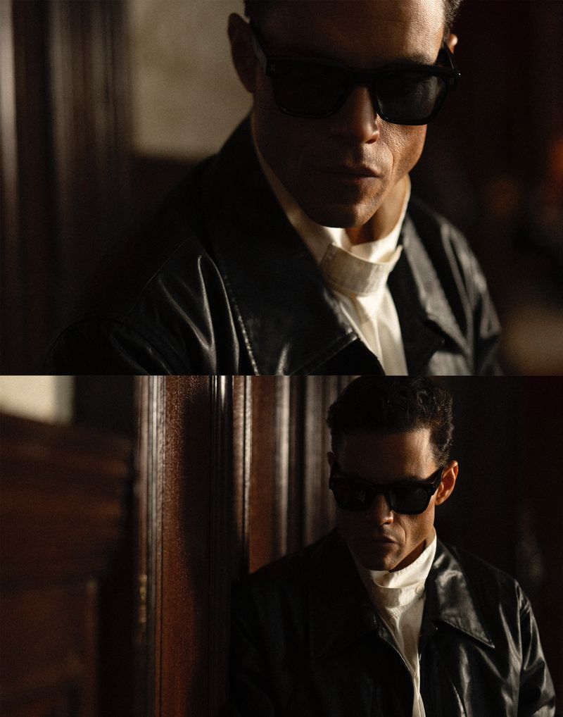 Rami Malek in dark sunglasses and a black leather jacket stands indoors with moody lighting and a neutral expression, looking down.