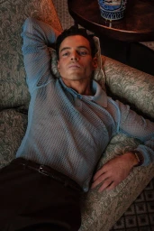 Rami Malek reclining on a patterned damask sofa wearing a sheer powder blue mesh knit polo shirt and dark trousers.