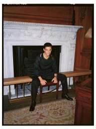 Actor Rami Malek sitting on a small chair in front of an ornate white stone fireplace in an interior room
