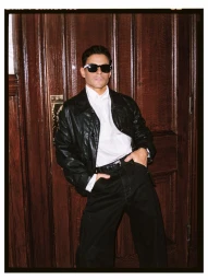 Actor Rami Malek posing in black sunglasses, a black leather jacket, white shirt, and black trousers against a wooden door.