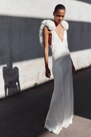 Model wearing a floor-length white plunging gown with textured shoulder trims against a sunlit wall casting linear shadows.